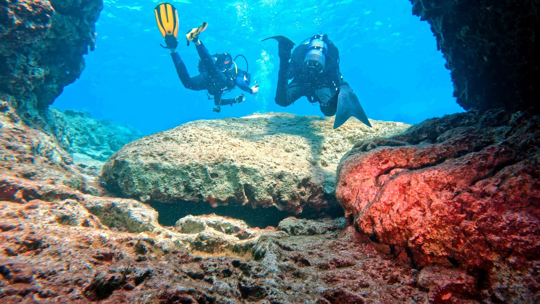 Two divers swimming through a rock arch in bright sunlit water