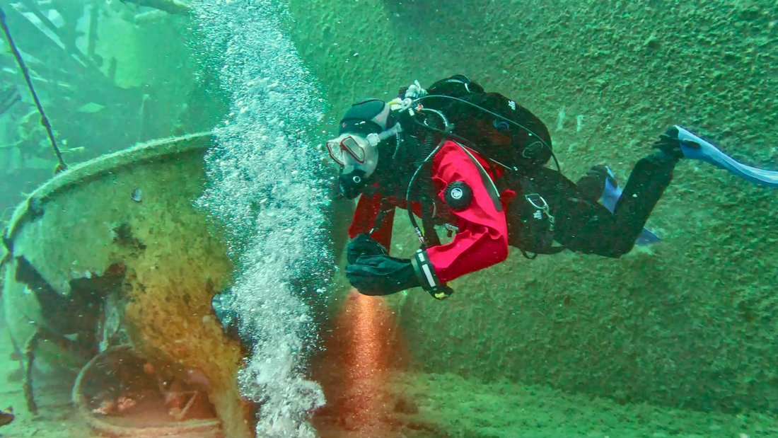 A diver in a red drysuit trailing bubbles over a wreck, the reds brought back