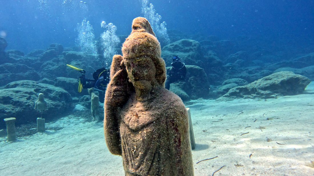 An underwater statue on the seabed with a diver behind, in clear blue water